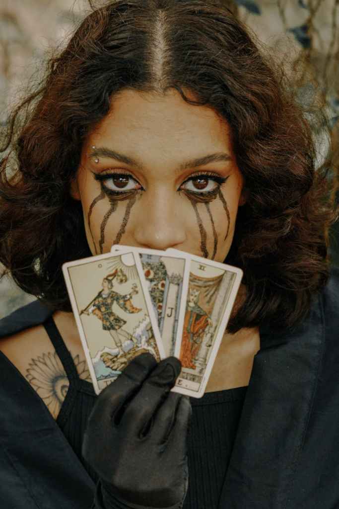 A person with dark, curly hair, wearing black gloves, holding tarot cards close to their face, with artistic tear makeup running down their cheeks.
