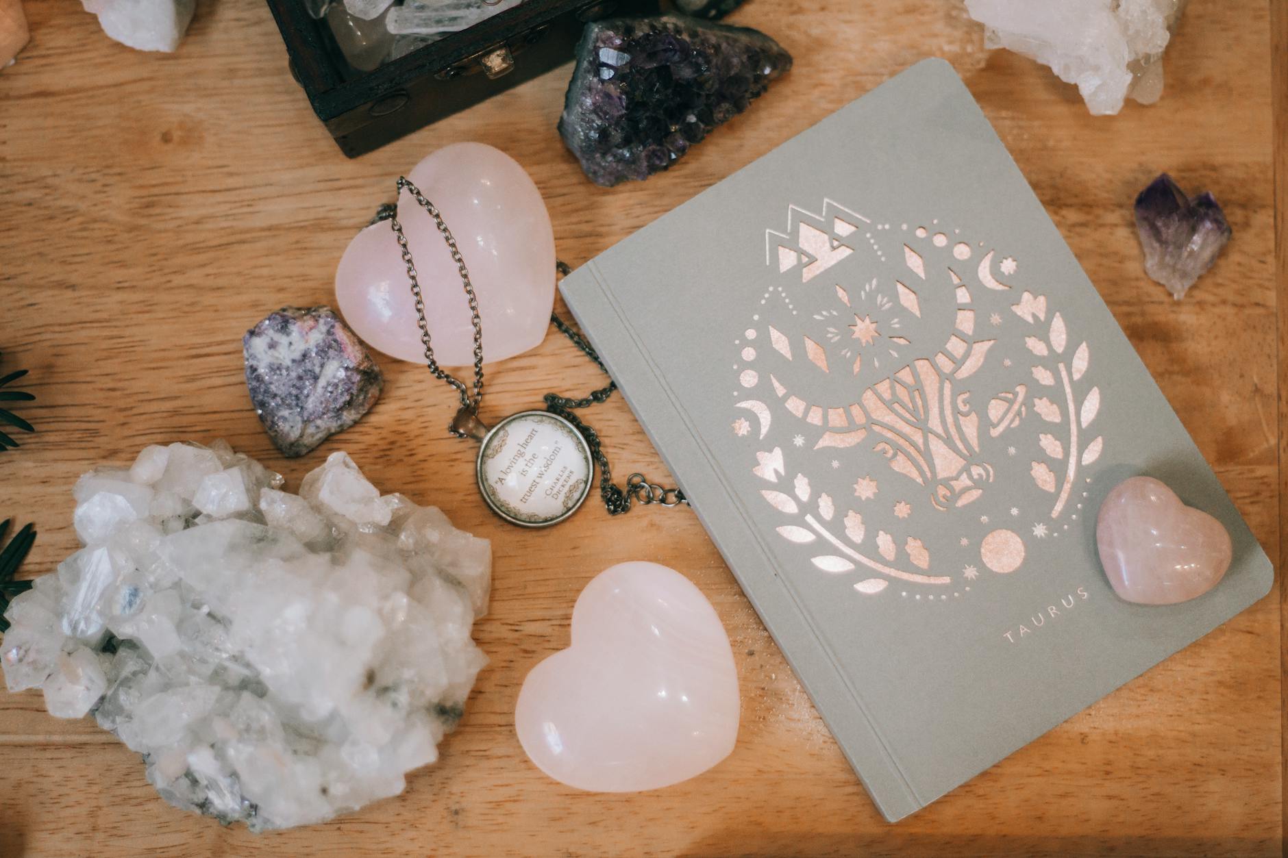 A collection of crystals and a notebook with a Taurus design on a wooden surface, surrounded by heart-shaped stones.
