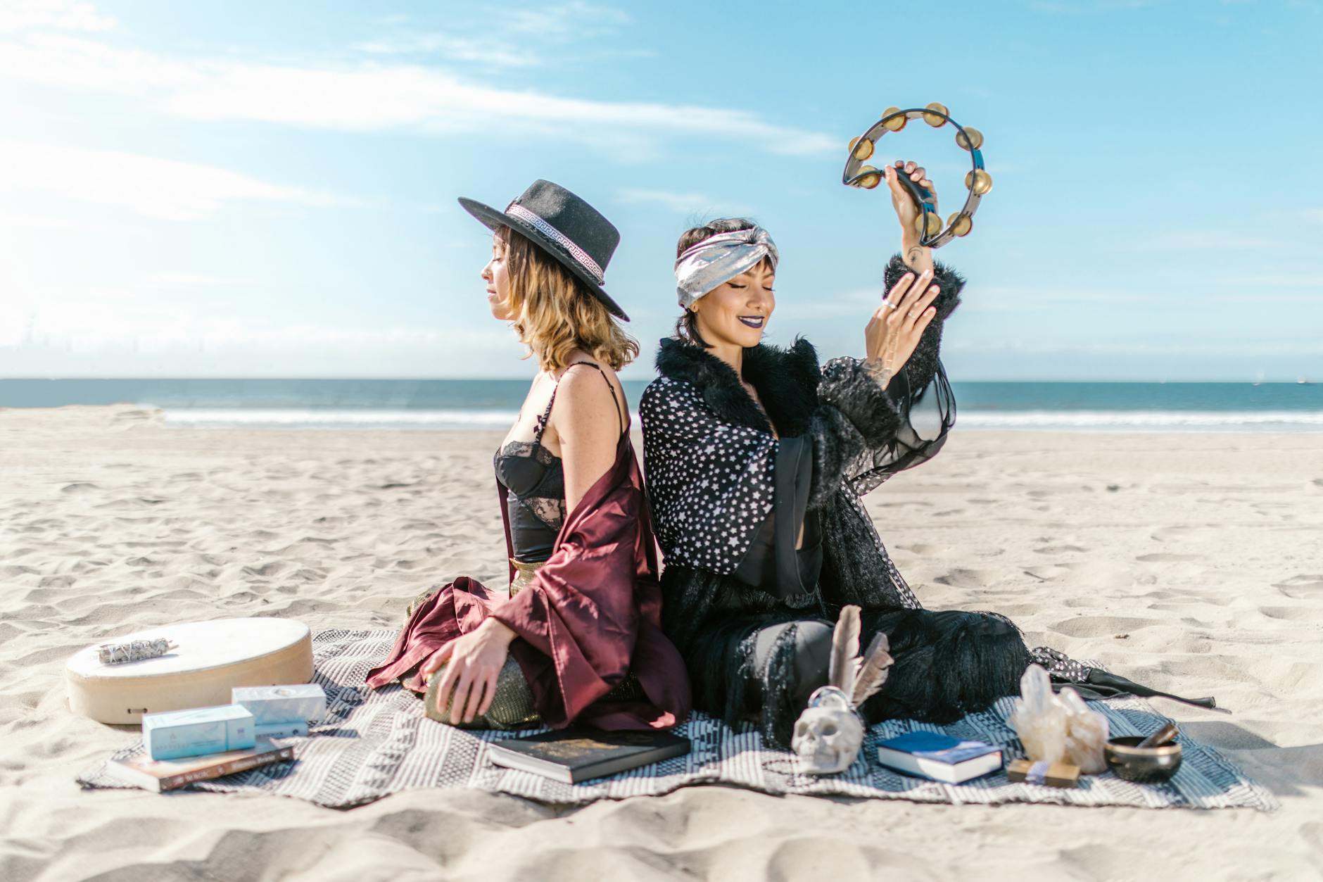 Two women sit back-to-back on a sandy beach, one wearing a black hat and the other a patterned scarf. They are surrounded by spiritual objects, including pillows, books, and a tambourine, with the ocean in the background.