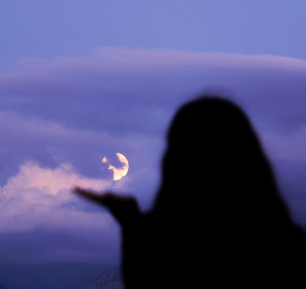Silhouette of a person reaching out towards a partially visible moon in a twilight sky filled with clouds.