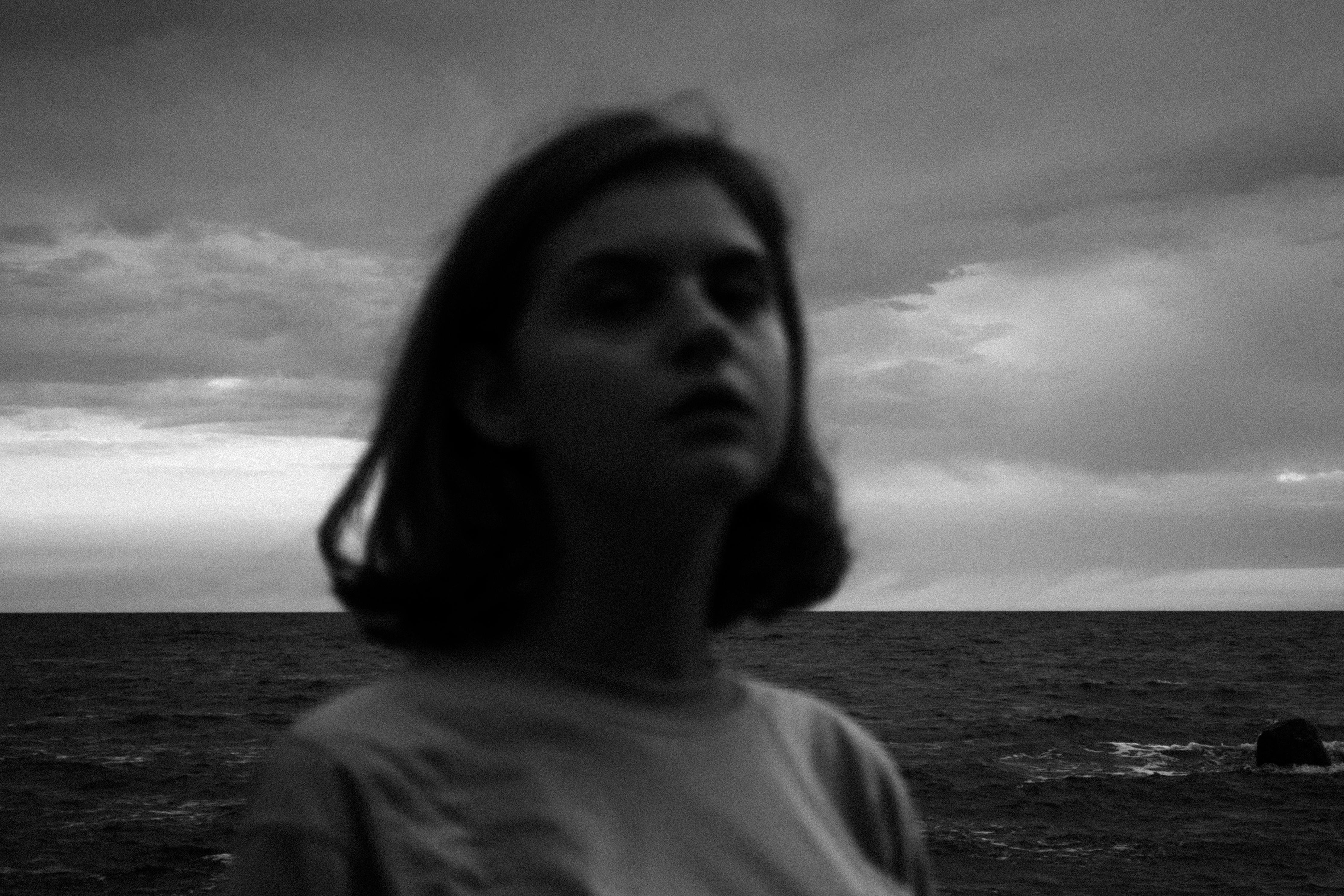 A blurred black-and-white portrait of a person standing near the ocean, with a cloudy sky in the background.